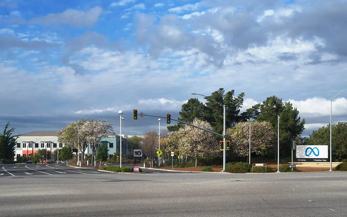 The entrance to Meta's headquarters in Menlo Park, California, with the Meta sign beside a traffic-light intersection under a cloudy sky