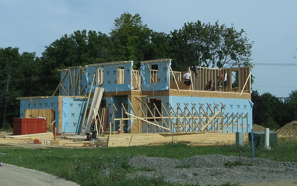A two-storey wood-framed house under construction on a bare lot in a Michigan subdivision, with workers on the upper floor and trees behind